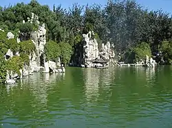 Rocky, bushy bluffs at the rear of some green water. There is a small structure with a Chinese roof at the base of one, in the center