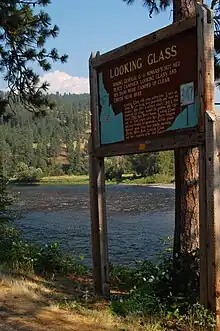 Color photograph of an explanatory panel in the foreground of a river flowing to the right, along a forest, under a blue sky.