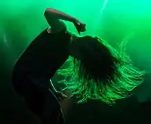 A picture of Lorde performing at a concert. Her body is captured in silhouette form with a green light in the background as she dances wildly. She bends her back forward as her hair moves forward with her movement.