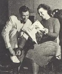 A smiling dark-haired woman holds up a copy of Chatelaine Magazine, which the man sitting beside her looks at as he ties his shoelaces.