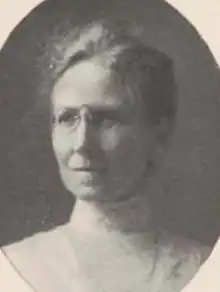 An older white woman with greying hair, wearing pince-nez glasses and a high-collored white blouse or dress