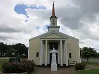 Photo shows a tan-colored church with the statue of a man in front.