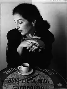 Black and white image of Fili sitting at table. Her hands are crossed over a cup of coffee and the table is decorated with Modernist type.