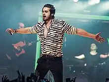 A dark haired man wearing a striped shirt extends his arms to the crowd while performing with a microphone