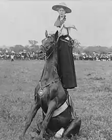 Lucille Mulhall, standing on the back of a seated horse