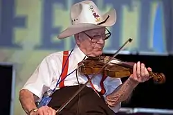 An elderly Luderin Darbone playing the fiddle. He is sitting and wearing a cowboy hat.