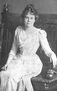 A white woman, seated in an ornate wooden chair. She is wearing a white corsetted gown. Her hair is arranged in an updo.