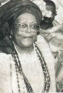 a black and white headshot of Mãe Cleusa Millet wearing a headdress, glasses, beaded necklaces, and white clothing