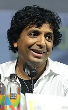 M. Night Shyamalan at a San Diego Comic-Con panel wearing a white shirt and talking into a microphone.