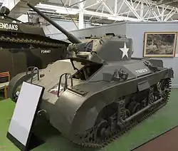 A squat dark green tank in a museum, placed on a large green felt mat. There is a description board in front of it, and a grey divided wall to its rear. The tank's gun barrel is pointing towards the ceiling and its left-hand caterpillar tracks are visible, as well as several large handles on its hull.