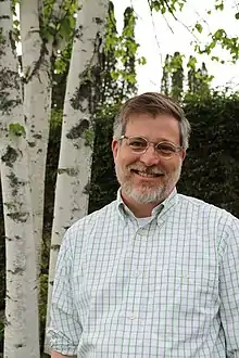 President Matthew Derr smiling and standing outside his home in Craftsbury Common, VT in front of a white paper birch tree and a green hemlock hedge