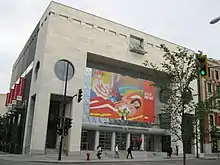 An almost cubical modernist building several stories high faced in smooth stone, with a recessed entrance on the ground level and circular windows above, seen at an angle from across the street. A large poster in front advertises a pop-art exhibit