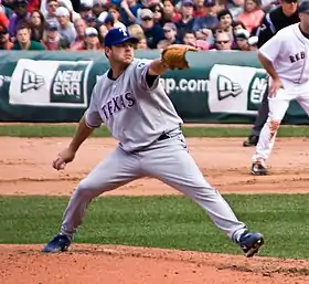 A man throwing a baseball with his right hand and wearing a gray baseball uniform with blue trim and "Texas" across the chest in block print and a blue baseball cap