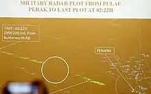 Brown background with white lines, dots, and labels depicting air routes, waypoints, and airports. Label in the top of the image reads: "Military radar plot from Pulau Perak to last plot at 02:22H." Green specks form a trail from bottom centre to left centre that was Flight 370. As the caption explains, the path is in two parts, with a white circle around the blank area between them and appears to highlight a section where the aircraft was not tracked by radar. Label at left end of flight path reads: "Time-02:22H 295R 200nm from Butterworth AB"