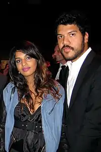 A man and a woman standing together. Both black hair and dark skin. The woman is wearing a light blue jacket over a black dress, and the man is wearing a white shirt and a black jacket.