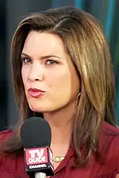 An image of a woman with shoulder-length hair and a dark red outfit. She is speaking into a microphone with the words "TV Guide".