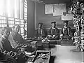 The police court. The magistrates are in overcoats and caps, with their clerks. Court records are fastened to walls and pillars.