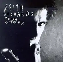 Black and white close-up photograph of Richards' face, brightly lit on right side of picture, hidden in shadow on left side, with KEITH RICHARDS MAIN OFFENDER in white scribbled capitals in top left quarter of album cover