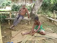 A Malagasy girls sits on the ground weaving with reeds, while an older gentleman sits on a bench above her.