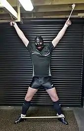 A male model is standing in a garage. He is stretched out into a standing spread eagle X shape and his wrists and ankles are secured with rope preventing him from moving freely.