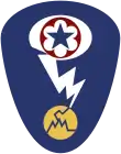 Oval shaped shoulder patch with a deep blue background. At the top is a red circle and blue star, the patch of the Army Service Forces. It is surrounded by a white oval, representing a mushroom cloud. Below it is a white lightning bolt cracking a yellow circle, representing an atom.