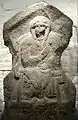 Gritstone bas-relief tombstone of Romano-British woman. There is a woman sculpted into the stone. She is seated in a high-backed chair. She has two plaits hanging down the front of her body and her right hand is poised towards her left shoulder.