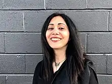 Woman with black hair, a black coat, and a silver necklace smiling into the camera, with a gray cinderblock wall in the background