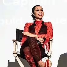 Performer Marawa the Amazing holds a microphone and smiles while sitting in a [[Director's chair]]. Her hair is pulled back and she is wearing a red outfit with red lips and long, dangling earrings.