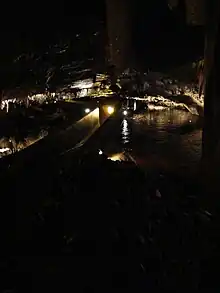 Cave passage with some features lit; within it a concrete pathway sinks to shoulder height with concrete walls either side. Dark brown water runs past outside and just below the top of the walls.