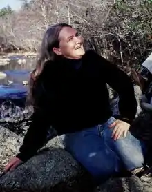 Photograph of a woman wearing jeans and a long-sleeved black sweater sitting on a river rock.