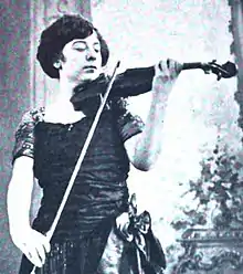 A young white woman with short dark hair playing a violin; she is wearing a dark dress with short sleeves