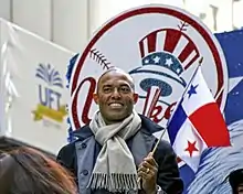 Mariano Rivera wearing a dark pea coat and gray scarf smiles while holding a red, white, and blue flag. He stands in front of a red, white, and blue logo that reads "Yankees".