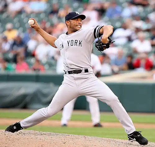 A right-handed Hispanic baseball pitcher, wearing a grey uniform with the lettering "NEW YORK" across it, with his body facing the right as he prepares to throw a baseball.