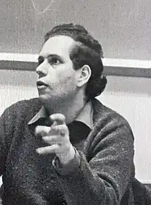 Photo of a man with dark hair gesturing in front of a classroom