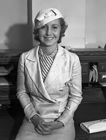 Martha Tilton sits atop a desk, wearing a light suit with a striped blouse.