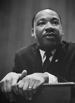 African-American man in his forties, dressed in a suit and tie, leaning and grasping on the edge of a lectern