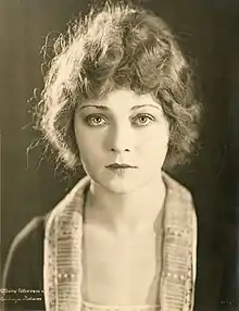 A sepia-toned photograph of a young white woman, her hair up off her collar )(either short or in an updo), wearing a shawl-collared jacket.