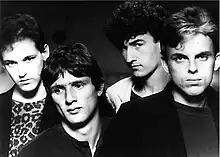 Head and shoulders shot of four men. Man at left has dark hair, face slight tilted to his right, but looking forward, he wears a dark jacket over a print tee-shirt. Second man is the shortest, also dark haired, his head is tilted further with eyes looking to his right, he wears a dark tee-shirt. Third man is the tallest, also dark haired, with his face partly in left profile, his head is also titled, his eyes are direct to the viewer, he wears a light coloured jacket with a dark tee-shirt. The last man is facing forward, he is the second tallest, his hair is lighter with fair coloured streaks at front, his jacket is dark over a less dark tee-shirt.