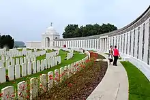 Colour photograph of the memorial stone wall and panels with war graves on grass shown to the right. There are red poppies growing in a flower bed to the left of the path and in front of the graves.