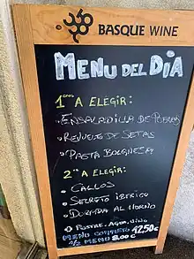 A board advertising "BASQUE WINE". It lists: MENU DEL DIA. 1eros A ELEGIR: · ENSALADILLA DE PUERROS · REVUELTO DE SETAS · PASTA BOLGNESA [sic] / 2os A ELEGIR: · CALLOS · SECRETO IBERICO · DORADA AL HORNO + POSTRE, AGUA, VINO / MENU COMPLETO 12,50 € / ½ MENU 8.00 €