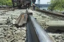 A view along the top surface of a rail with another piece of rail twisted around it a short distance away and a freight car on the track behind it. The ties and ballast in the foreground are a much lighter color than those elsewhere in the image