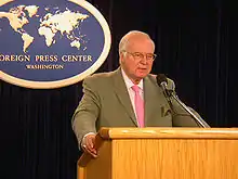 A white-haired man in a suit and pink tie is standing at a lectern, speaking into a microphone. Behind him is a plaque reading "FOREIGN PRESS CENTER WASHINGTON".