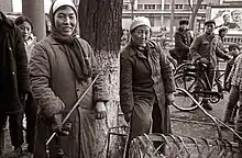 A black and white photo of two people in heavy coats, scarves, and hats holding tools smile candidly at the camera. Several others look at the camera from the background.