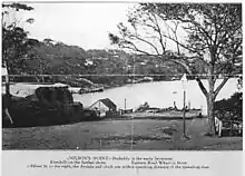 Postcard of Milsons Point. The caption reads: "Milson’s Point – Probably in the early Seventies [1870s]. Kirribilli on the farther shore. Eastern Road Wharf is front. Alfred St to the right, the Arcade and clock are within (illegible)ing distance of the spreading tree."