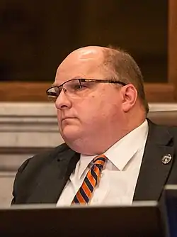 A bespectacled man in a black blazer and orange-and-purple striped tie sits behind a computer.