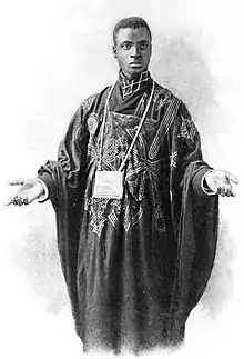 A black man, standing, with close-cropped hair and hands stretched forward and outward. He is wearing heavy embroidered robes.