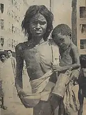  A very shriveled and emaciated woman holding a very emaciated baby in the crook of her arm. Both are wearing only rags, and the mother's right breast is unconcealed. Her hands are holding a bowl and a canister, and she may be begging.