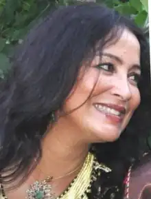 Color portrait photo of a dark-haired, smiling woman wearing a multi-colored top and a jeweled choker necklace.