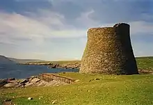 Green fields lie to the right of a body of dark blue water. At the water's edge, there is a tall, round, stone tower, possibly unroofed.