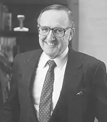 A smiling man with pale skin, glasses and wearing a suit with a spotted tie